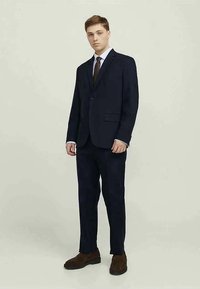 Fato azul-marinho com um casaco de botão único, lapelas recortadas e bolsos laterais. Combinado com uma camisa branca, gravata escura e sapatos castanhos.