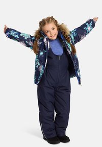 Blå blommig puffjacka med pälsdetalj, marinblå overaller och svarta stövlar. Barnet poserar med utsträckta armar och visar upp outfitens design.