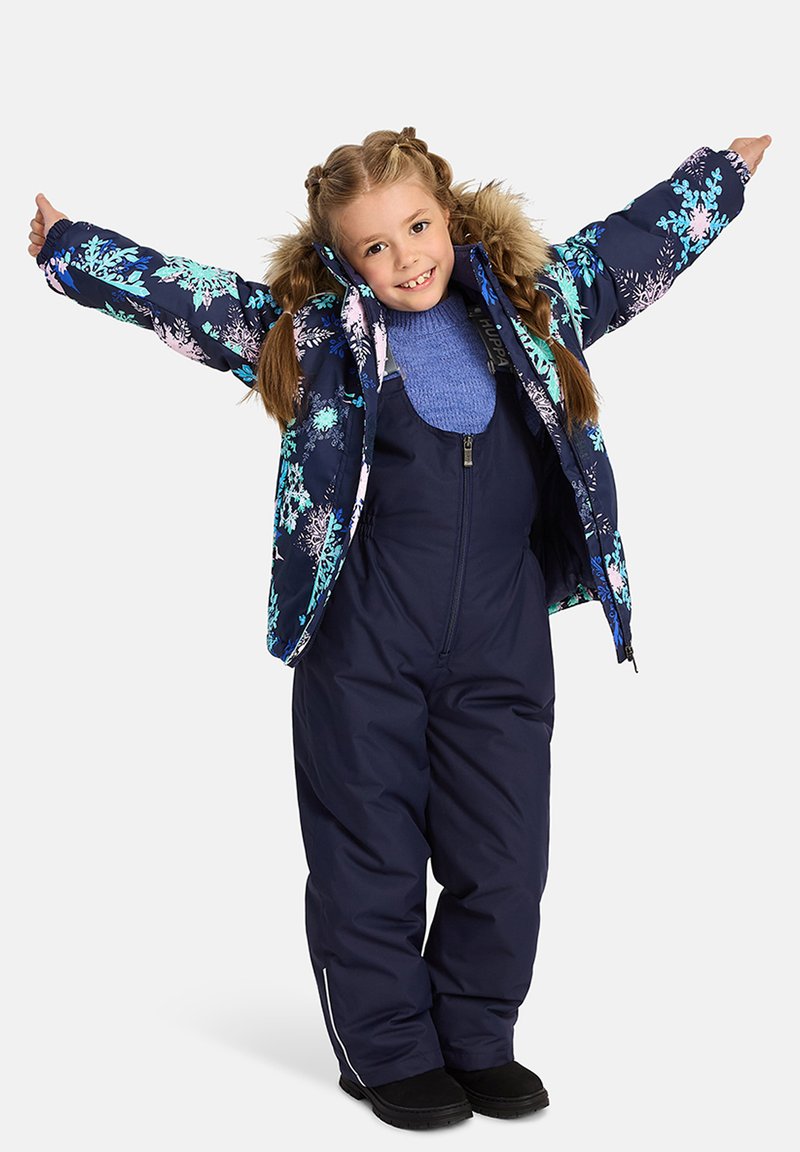 Blå blommig puffjacka med pälsdetalj, marinblå overaller och svarta stövlar. Barnet poserar med utsträckta armar och visar upp outfitens design.