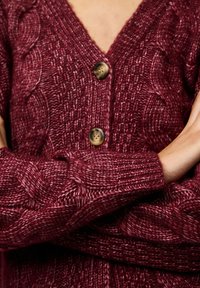 Burgundy stickad kofta med V-ringning, som har ett kabelmönster, träknappar och ribbade ärmslut, med en strukturerad design.