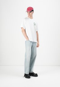Witte oversized t-shirt met groene print, lichtblauwe rechtuitlopende jeans en zwarte chunky schoenen. Rode pet met zwarte accenten maakt de look compleet.