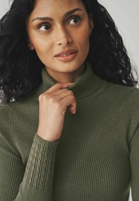 Pull en maille côtelée vert olive avec des accents texturés sur les manches. Gros plan sur le col et une main reposant près du menton, mettant en valeur le détail du tissu.