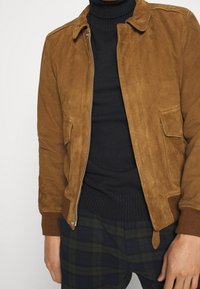 Brun mockaläderjacka med dragkedja, spetsig krage, två framfickor och ribbade ärmslut. Bärs över en svart ribbad polotröja.