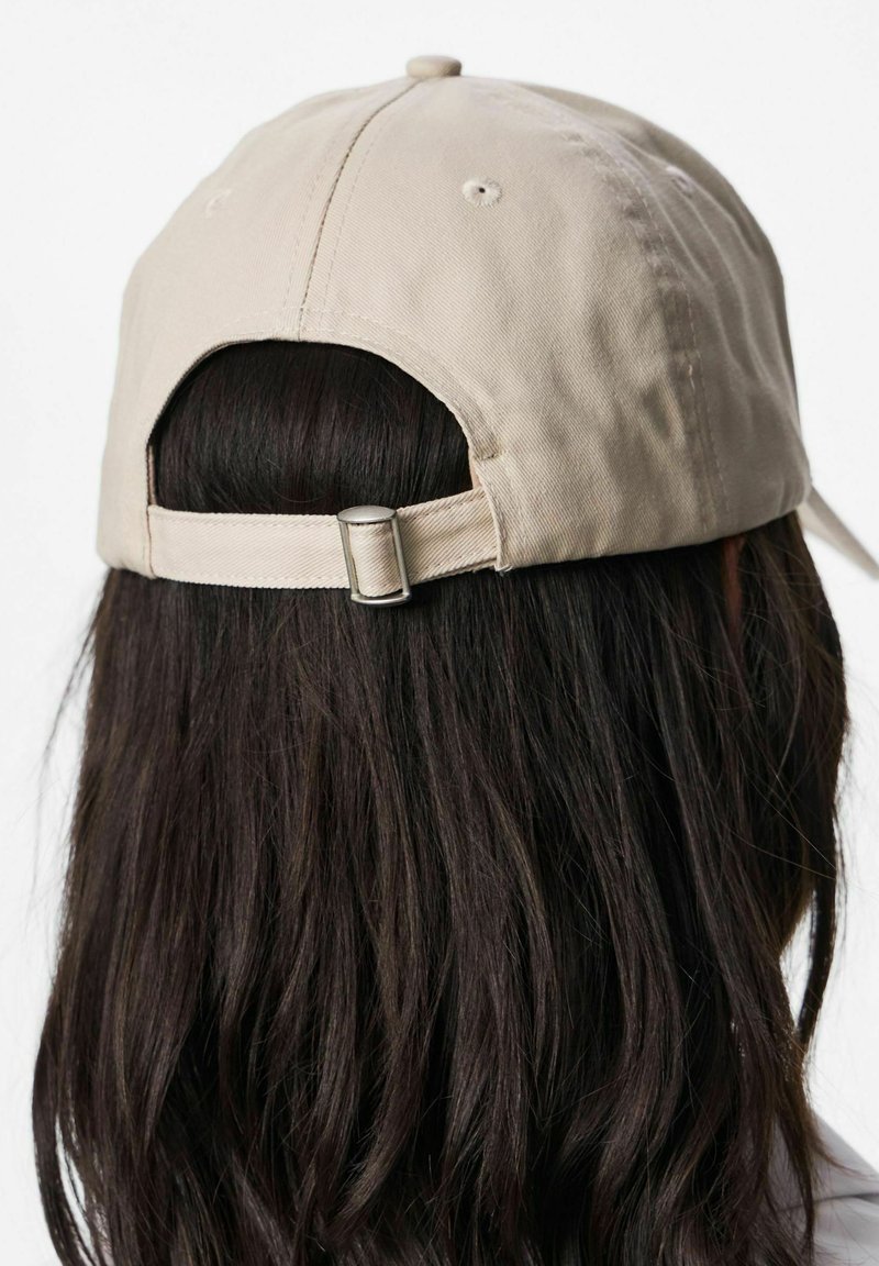 Femme aux longs cheveux foncés portant une casquette de baseball beige vue de dos, montrant la sangle réglable et la boucle métallique.