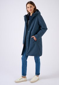 Cappotto impermeabile blu navy con cappuccio, zip frontale e bottoni a pressione. Presenta due tasche laterali e una vestibilità ampia, lunga fino al ginocchio.