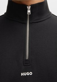 Felpa nera con zip e collo alto, con una cerniera argentata e un testo del logo bianco, realizzata in un tessuto morbido e strutturato.