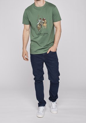 Grön bomullströja med en tryckt design av polospelare på hästryggen; matchad med mörka jeans och vita sneakers.