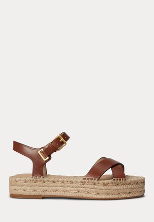 Sandale maro din piele cu bretele încrucișate, cataramă la gleznă și talpă groasă din iută țesută, pe un fundal deschis simplu.