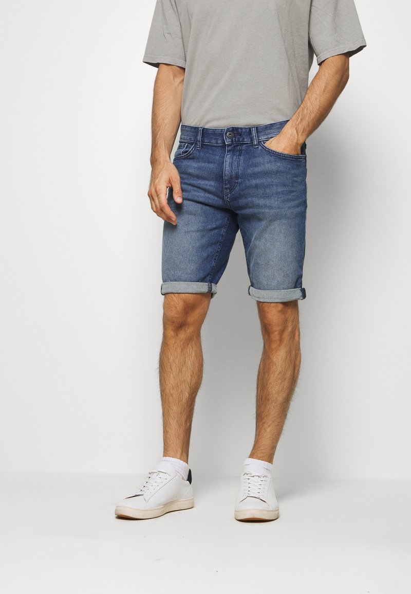 Homme portant un short en denim bleu retroussé, un T-shirt gris, des chaussettes blanches et des baskets blanches, debout devant un fond clair uni.