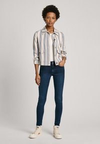 Chaqueta de algodón a rayas en beige con rayas azul y navy, con bolsillos en el pecho y un diseño entallado, combinada con vaqueros oscuros ajustados y zapatillas.