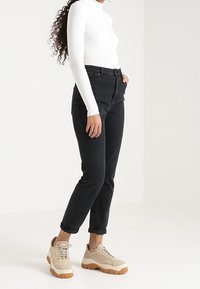 Schwarze, hoch taillierte Jeans mit schmalem Bein, kombiniert mit einem weißen, gerippten Langarmoberteil und beigefarbenen Sneakers mit strukturiertem Profil.