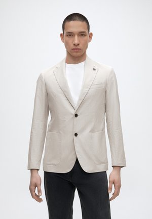 Giovane uomo in piedi su sfondo bianco che indossa un blazer beige chiaro sopra una camicia bianca e pantaloni neri.