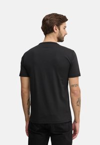 Camiseta negra de manga corta con cuello redondo, textura suave y corte relajado, vista desde atrás. Tatuaje visible en el brazo.
