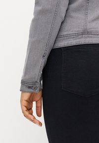 Manga de chaqueta de mezclilla gris con un detalle de botón, combinada con tela negra. La manga está ligeramente doblada, mostrando la costura y la textura.