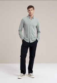 Cămașă lungă cu mâneci verzi deschise, cu guler cu nasturi, asociată cu pantaloni bleumarin. Modelul poartă adidași gri. Design minimalist, textură netedă.