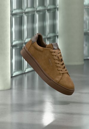 Brauner Wildleder-Sneaker mit passenden Schnürsenkeln und Sohle, mit dem Brand "GANT" an der Seite, vor einer Glasbausteinwand und poliertem Boden.
