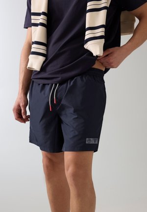 Mężczyzna noszący granatowe szorty kąpielowe ze sznurkiem, granatową koszulkę oraz kremowy sweter z granatowymi paskami zarzucony na ramiona.