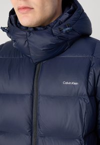 Navy puffer jacket with a high collar, zipper closure, and Calvin Klein logo. The fabric has a smooth, glossy texture and quilted design.