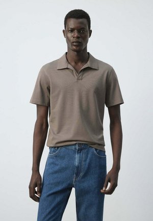 Hombre de pie con una camiseta tipo polo marrón de manga corta y vaqueros azules sobre un fondo blanco liso.