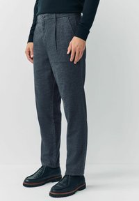 Mężczyzna w ciemnoszarych, wzorzystych spodniach, czarnych skórzanych butach na sznurówki i ciemnej, długiej bluzie, stojący na tle jasnego, jednolitego tła.