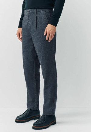 Muž oblečený v tmavě šedých texturovaných kalhotách, černých kožených šněrovacích botách a tmavém topu s dlouhým rukávem, stojící na jednoduchém světlém pozadí.