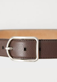 Brun lærrem med en rektangulær sølvspenne, med små firkantede utsparinger og sømdetaljer, merket med "Calvin Klein."