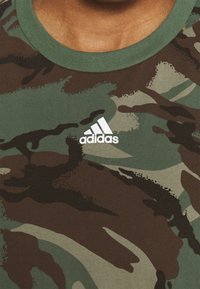 Zelené a hnědé maskáčové tričko s designem kulatého výstřihu; obsahuje bílé logo Adidas na hrudi. Měkká, strukturovaná tkanina.