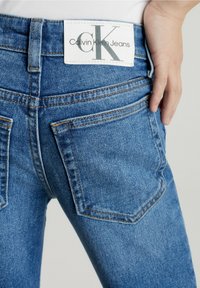 Los jeans de mezclilla azul presentan una cintura alta, bolsillos traseros y una etiqueta blanca con "Calvin Klein Jeans" en letras grises.