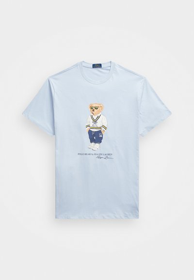 Himmelblå bomullst-skjorte med grafikk av en bjørn med solbriller og en hvit sportstrøye med blå og gule aksenter. Teksten under leser "Polo Bear av Ralph Lauren."
