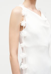 Bezrokavu balta kleita ar caurspīdīgām tīkla malām, rotātām ar mazām 3D ziedu aplikācijām, valkāta uz augšējās ķermeņa daļas un pleca.
