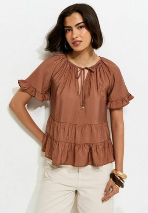 Femme portant un chemisier marron à manches courtes avec des volants et un col noué, ainsi qu'un pantalon beige, accessoirisée avec de grands bracelets et des boucles d'oreilles créoles.
