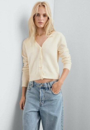 Gilet court en crème clair, avec un décolleté en V, des manches longues et une fermeture à boutons. Associé à un jean taille haute bleu clair. Texture douce.