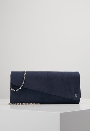 Pochette en daim bleu marine avec fermeture à rabat et bandoulière chaîne argentée, reposant sur une surface géométrique blanche sur fond gris.