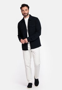 Veste en polaire noire avec deux poches poitrine, associée à une chemise blanche et un pantalon clair, portée avec des chaussures sombres. Design simple et moderne.