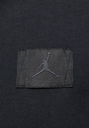 Mustan kankaan päällä on ommeltu suorakaiteen muotoinen läntti, jossa on kohotettu siluetti koripalloilijasta hyppäämässä pallon kanssa.