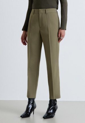 Donna con pantaloni verde oliva su misura, maglia a maniche lunghe verde scuro e stivaletti neri con tacco alto e punta, in piedi su pavimento grigio.