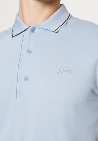 Světle modrá polo košile vyrobená z bavlny s límcem, která má dva knoflíky, černé lemování na límci a vyšívané logo "BOSS".