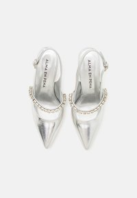 Sandały na obcasie w metalicznej srebrze, z ostrym noskiem i ozdobnym łańcuszkiem na górze. Gładka tekstura i otwarty design.