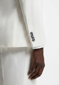 Mano de piel oscura junto a la manga de un traje de lino blanco con cuatro botones oscuros en el puño, pantalones blancos a juego visibles con iluminación suave.