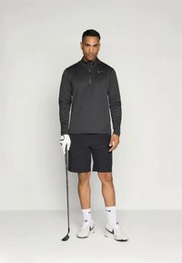 Μαύρο μακρυμάνικο performance top με φερμουάρ, λογότυπο Nike, συνδυασμένο με σκούρα σορτς, λευκές κάλτσες και αθλητικά παπούτσια, κρατώντας ένα κλαμπ γκολφ.