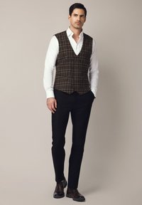 Brązowa kamizelka w kratę z dekoltem w serek i zapięciem na guziki, noszona na białej koszuli, w połączeniu z czarnymi spodniami i brązowymi skórzanymi butami.