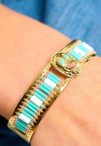 Bracelet en or avec des perles turquoises et blanches, présentant un motif tissé complexe. Décoré d'un détail central en pierre précieuse.