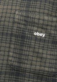 Flisasta srajca s karo vzorcem v sivi in bež barvi, ki ima žep na prsih in bele vezenine "obey" na žepu.