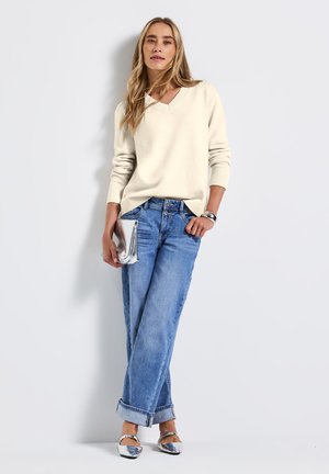 Hellbeige Strickpullover mit V-Ausschnitt, kombiniert mit hellblauen weiten Jeans und silbernen Ballerinas. Hält eine kleine silberne Clutch.