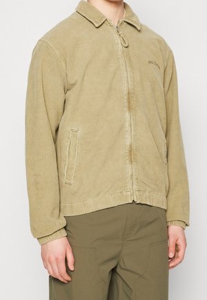 Homme portant une veste zippée en velours côtelé beige et un pantalon vert olive, debout devant un fond clair uni.