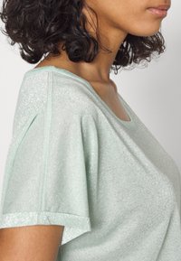 Femme aux cheveux courts et bouclés portant un haut à manches courtes scintillant vert clair, vue de profil au niveau de l'épaule et du cou.