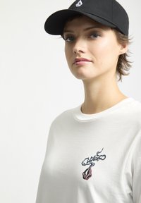 Hvid bomulds t-shirt med rund halsudskæring, prydet med et farverigt broderet design på brystet, sammen med en sort kasket.