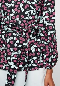 Top in tessuto nero con un motivo di ciliegie e foglie in rosa e azzurro chiaro. Include una cintura da annodare in vita e maniche a sbuffo per un dettaglio in più.