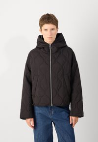 Veste matelassée noire avec capuche, dotée d'une fermeture éclair centrale, de poches latérales et d'un motif en vagues texturé. Porte avec un jean bleu.