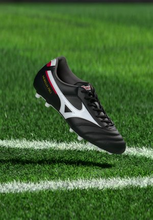 Μαύρο και άσπρο ποδοσφαιρικό παπούτσι Mizuno Morelia Pro που αιωρείται πάνω από πράσινο χορτάρι κοντά σε μια λευκή γραμμή γηπέδου.